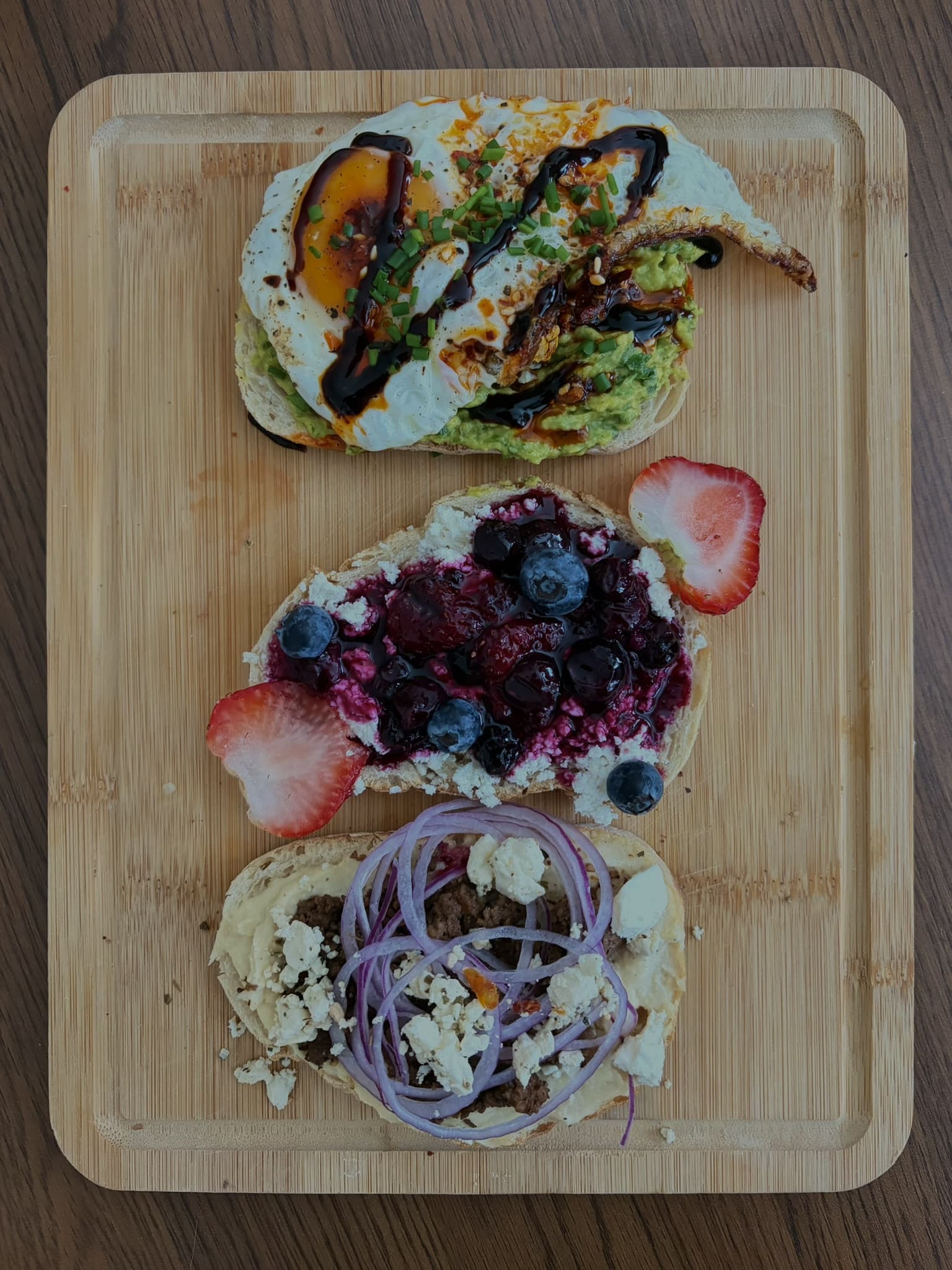 Three open-faced toasts on a bamboo board: avocado with fried egg, honey feta with berries, and beef with hummus
